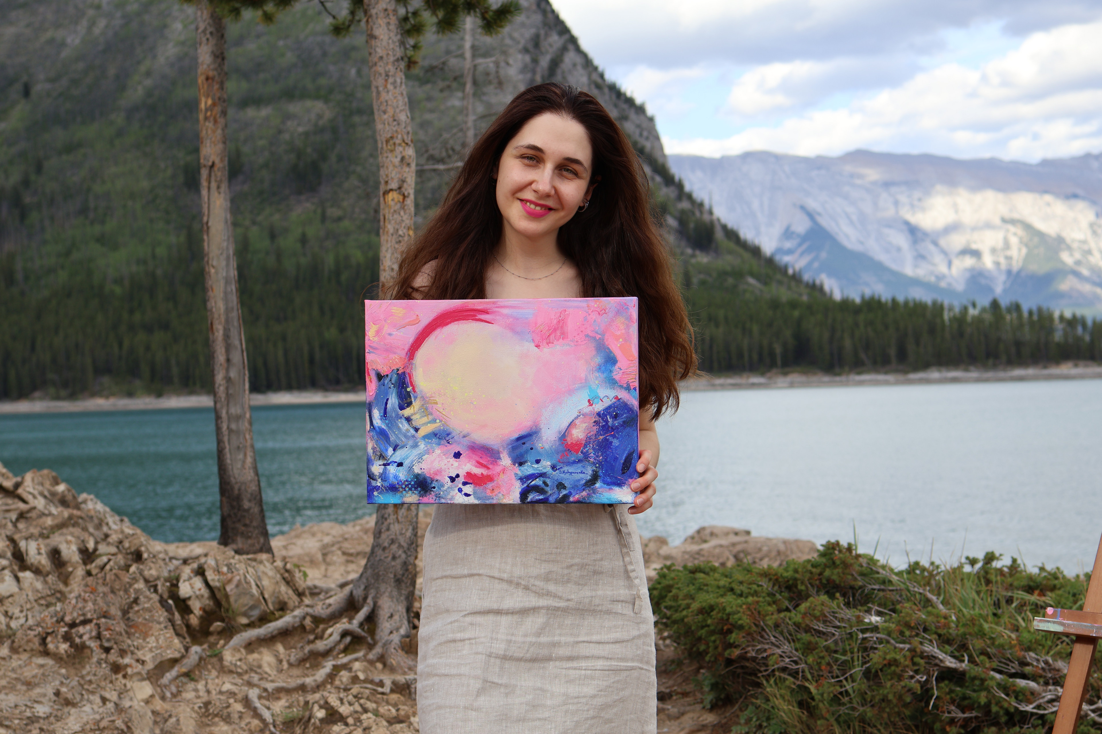Woman holding a colorful painting outdoors with a scenic background of trees and mountains.